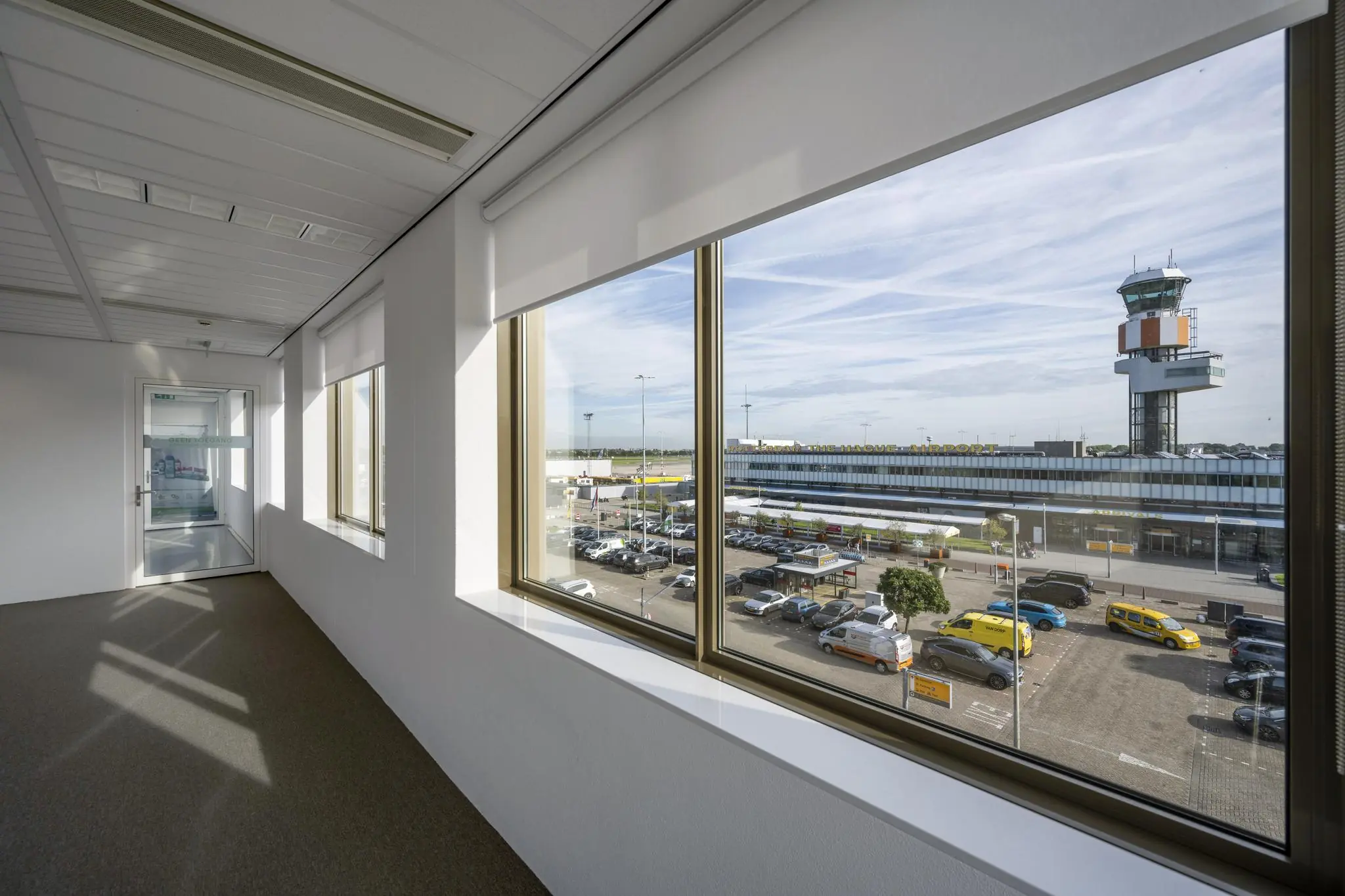Uitzicht vanuit een gang op Rotterdam The Hague Airport met parkeergelegenheid en de verkeerstoren op de achtergrond.